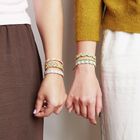 Zwei Hände, die bunte Freundschaftsarmbänder tragen, halten sich. Sie tragen bunte Oberteile.