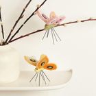 Zwei Filzschmetterlinge, einer rosa, einer orange, mit Drahtbeinen, auf Zweigen und einer weißen Vase.