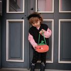 Ein Kind in schwarz-weiß gestreiftem Pullover mit Kürbistasche und Spinnen-Haarreif steht vor einer grauen Tür.