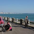 Nummernschild: 1434 RD 76. Eine Frau wechselt am roten Auto den Reifen, ein Mann in Mantel schaut aufs Meer.