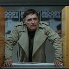 "MOODY WHITE STAR" auf Postern im Hintergrund. Ein Mann im beigefarbenen Trenchcoat zwischen zwei Automaten.