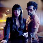 Eine Frau in einem schwarzen Outfit hält ein Papier und spricht mit einem Mann an einer Bar. Dämmerige, farbige Beleuchtung.