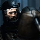 Zwei Menschen in dunklen Schutzanzügen stehen nah beieinander; der vordere ist unscharf, der hintere zeigt ein ernstes Gesicht.