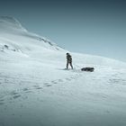 Eine Person zieht einen Schlitten durch eine verschneite, bergige Landschaft unter klarem Himmel.