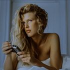 Eine Frau mit langen, lockigen Haaren hält ein Mobiltelefon im Bett, mit einem nachdenklichen Ausdruck und umgeben von eleganten Wänden.