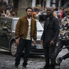 Zwei Männer stehen auf einer Kopfsteinpflasterstraße vor einem schwarzen Auto mit dem Kennzeichen "SY62". Im Hintergrund sind Menschen.