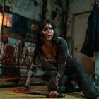 Eine Frau kniet auf einem blutverschmierten Boden, mit Blut im Gesicht und auf der Kleidung in einem verwahrlosten Raum.