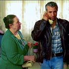 Eine ältere Frau und ein Mann mit Telefon. Frau trägt grüne Strickjacke, Mann hat Lederjacke. Im Hintergrund Vorhänge.