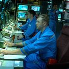 Männer in blauen Overalls arbeiten an Konsolen mit Bildschirmen in einem Kontrollraum voller Computer und Technik.