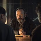 Ein Mann mit Brille und Bart sitzt an einem Tisch und spricht mit zwei Personen. Er hat die Hände gefaltet.