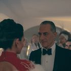 Ein Mann im Smoking, lächelnd, spricht mit einer Frau in einem eleganten roten Kleid auf einer formellen Veranstaltung.