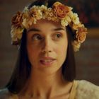 Eine Frau mit Blumenkranz im Haar, schaut leicht zur Seite, sanfter Gesichtsausdruck, unscharfer Hintergrund.