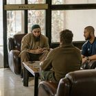 Text: "PULL". Drei Männer sitzen in einem Raum mit Ledersesseln an einem Holztisch. Einer trägt eine grüne Mütze.