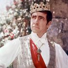 Ein Mann mit Krone und rotem Band steht vor einer Steinmauer. Im Hintergrund sind verschwommene Blumen zu sehen.