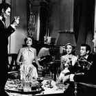 Fünf elegant gekleidete Menschen bei einem feinen Cocktailabend, sitzen und stehen in einem stilvollen Raum mit Spiegel und Lampe.