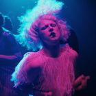 Eine Person mit weißer Perücke tanzt intensiv in blau-pinkem Licht, umgeben von Schatten anderer Menschen.