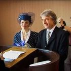 Ein Paar steht vor einem Pult; sie tragen elegante, festliche Kleidung. Ein Fotograf ist im Hintergrund zu sehen.