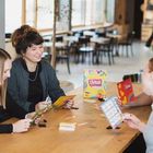 Vier Personen spielen fröhlich Brettspiele an einem Tisch in einem modernen Café. Im Vordergrund sind Spielkarten.