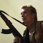 Ein blonder Mann mit schmutzigem Gesicht und brauner Weste hält ein Gewehr. Sein Blick ist entschlossen.