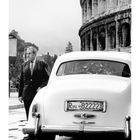 "Roma 82222X" auf Autokennzeichen, Mann im Anzug, Kolosseum im Hintergrund, Kalender zeigt Monat Juli.