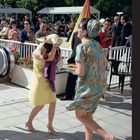 Zwei elegant gekleidete Frauen in Sommerkleidung, umgeben von Zuschauern. Im Hintergrund grüne Bäume und ein Pavillon.