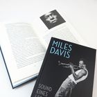 Buchcover mit Titel „Miles Davis: Sound eines Lebens“. Foto von Miles Davis mit Trompete. Ein geöffnetes Buch liegt daneben.
