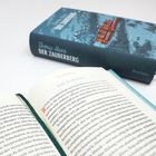 Ein geöffnetes Buch zeigt den Text "Fülle des Wohllauts". Im Hintergrund liegt ein geschlossenes Buch mit dem Titel "DER ZAUBERBERG".