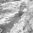 Keine erkennbaren Texte. Schwarz-weiß-Aufnahme eines verschmutzten Flusses mit kiesigem Ufer, Schaum auf dem Wasser.