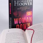 Der Text auf dem Buch: "Er soll sie retten. Aber seine Nähe bedroht alles, was sie hat. WOMAN DOWN". Ein aufgeschlagenes Buch mit Leseband.