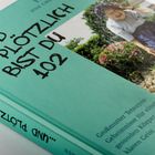 Auf einem mintgrünen Buch steht der Titel "...UND PLÖTZLICH BIST DU 102". Rechts ist ein Foto einer älteren Frau in einem Garten.