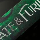"FATE & FURIES" steht in silbernen Buchstaben auf einem grün strukturierten Hintergrund.
