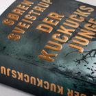 Goldene Schrift: "SØREN SVEISTRUP", "DER KUCKUCKSJUNGE", "THRILLER". Dunkles Cover mit kahlen Ästen im Hintergrund.