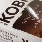 Text: "MICHAEL KOBI, SCHATTEN ÜBER SØMARKEN." Buchcover mit einem stimmungsvollen Himmel und einer Windmühle.