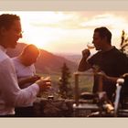 Kein Text vorhanden. Vier Männer genießen Getränke und Snacks bei Sonnenuntergang auf einer Terrasse mit Bergblick.