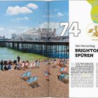 "74, Den Herzschlag Brightons spüren. Menschen am Kiesstrand mit bunten Liegestühlen, Brighton Pier im Hintergrund, blaues Meer."