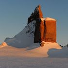 Verschneite Landschaft mit einem hohen, markanten Berggipfel im Abendlicht, der orangefarben leuchtet.