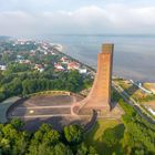 Roter Turm am Wasser, umgeben von üppigem Grün, rechter Hand eine Stadt, im Hintergrund das Meer.