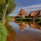 Ein altes Gebäude mit rotem Ziegeldach spiegelt sich in einem klaren Fluss, umgeben von grünen Bäumen und Wiese.