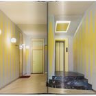 Keine sichtbaren Texte. Ein Flur mit gelb-grauen Wänden, runden Wandlampen, einem Aufzug und einer Treppe mit Marmorfliesen.