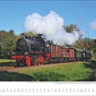 Juli: Dampflok mit Güterwaggons auf Schiene, rauchender Schornstein, umgeben von grüner Landschaft und blauem Himmel.