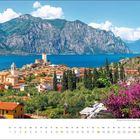Kalender für Juli, italienische Stadt am See mit Burg, Bergen im Hintergrund und lilafarbenen Blumen im Vordergrund.