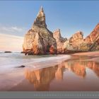 Text: "07 | JULI". Eine felsige Küste mit spitzen Felsen, ruhiges Wasser und einem sandigen Strand bei Sonnenuntergang.