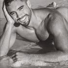 07. Ein lächelnder Mann liegt am Strand im seichten Wasser, sein Kopf wird von einer Hand gestützt.