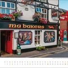 „ma bakers“ auf einem Pub-Schild, rote Fassade, hängende Blumen, Wandgemälde mit Menschen. Kalender unten: Juli.