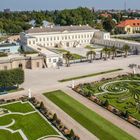 Prunkvolle Gartenanlage mit geometrischen Mustern, gepflegten Grünflächen und einem großen, weißen Gebäude im Hintergrund.