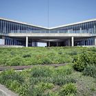 Moderner, winkelförmiger Betonbau mit vielen Glasfassaden, im Vordergrund grüne Vegetation.