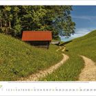 Kalenderseite für Juli. Ein Feldweg führt an einer Hütte mit rotem Dach vorbei, umgeben von grünen Wiesen und Bäumen.
