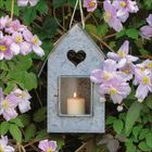 Metalllaterne mit Herzfenster, darin eine brennende Kerze. Umgeben von rosa Blumen und grünen Blättern.