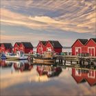 Sechs rote Holzhäuser mit schwarzen Dächern stehen am Wasser, im Vordergrund liegen Boote am Steg, der Himmel ist bewölkt.