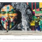 Das Wandgemälde zeigt ein Gesicht mit bunter, geometrischer Musterung auf einer Backsteinoberfläche. Unten ein Kalender.